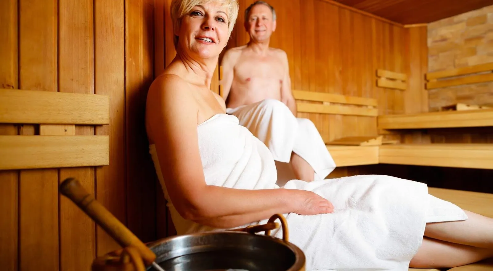 A smiling woman and a man wrapped in towels on the benches of a wooden sauna