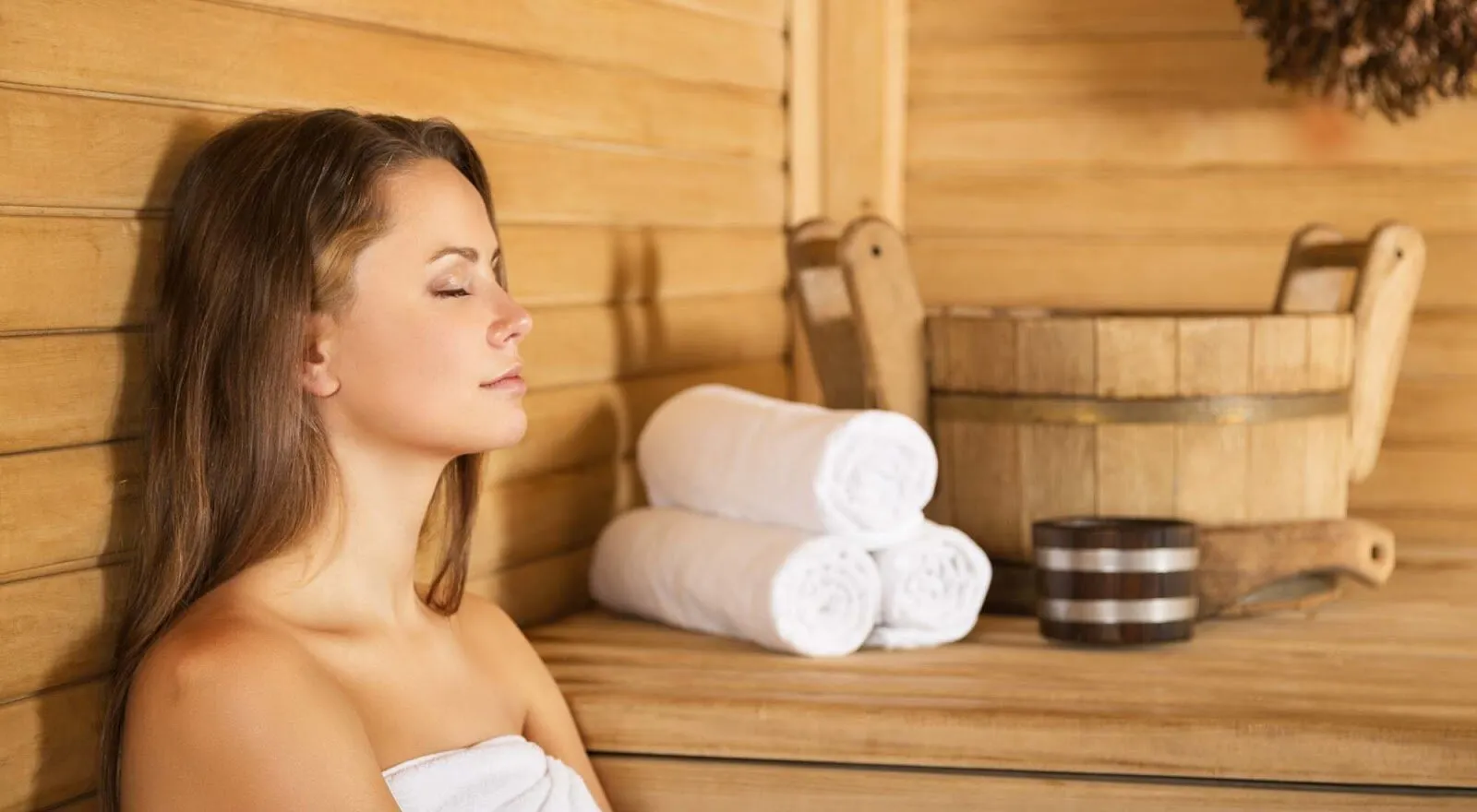 A woman resting with her eyes closed in a wooden sauna beside rolled towels and a sauna bucket