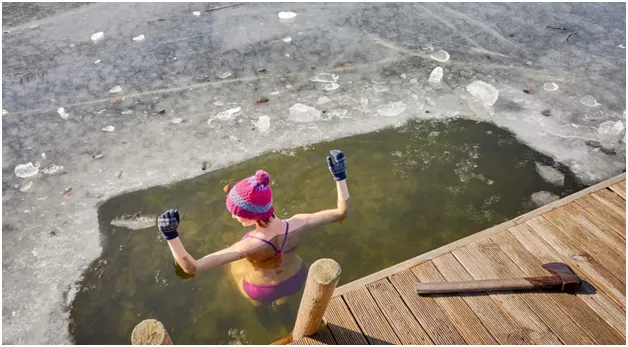 A person in a knit hat and gloves raising both arms in an icy plunge hole beside a wooden dock