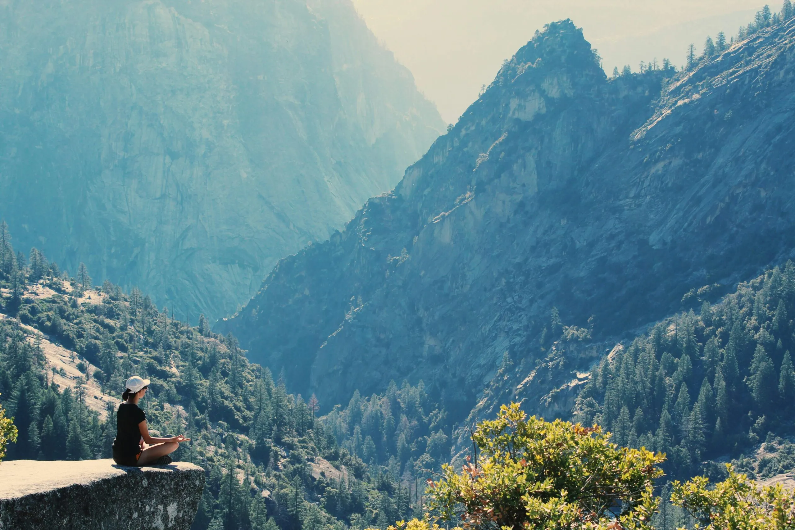 A person sitting cross-legged on a rocky ledge, looking out over a forested mountain valley.